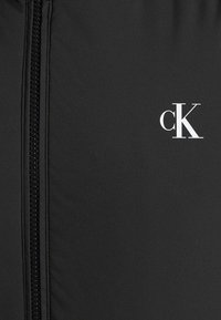 Veste zippée noire avec une texture lisse. Présente un logo "cK" blanc à l'avant et une fermeture éclair robuste sur toute sa longueur.