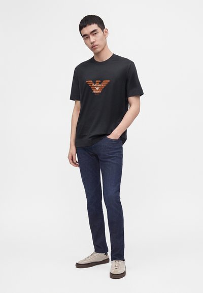 Νέος άνδρας που φοράει μαύρο T-shirt Emporio Armani, σκούρο μπλε τζιν και μπεζ αθλητικά παπούτσια, στέκεται με το ένα χέρι στην τσέπη.