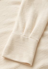 Tessuto beige con texture e design a righe. Il polsino è ripiegato, presentando sottili variazioni di tonalità e una finitura morbida e leggera.