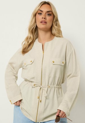 Blonde woman wearing a beige zip-up jacket with drawstring waist and gold button accents, holding sunglasses in one hand.