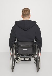 Felpa nera con cappuccio e cordino, seduta in una sedia a rotelle con uno schienale in tessuto nero e ruote argentate. Sneakers bianche visibili sotto.