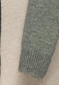 Neulottu villapaita, jossa on vaaleanbeige vartalo, kontrastiväriset tummanvihreät hihat, resorit hihansuissa ja pehmeä, teksturoitu pinta.