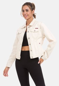 Witte denimjas met een klassiek ontwerp, voorzien van twee voorzakken, metalen knopen en contrasterende stiksels. Model draagt een zwarte top eronder.