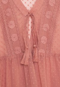 Hellrosa, strukturiertes Kleid mit floralen Spitzenakzenten und Quasten am Ausschnitt. Plissee-Stoff mit einem gepunkteten Muster.