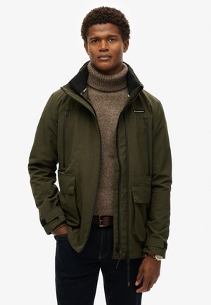 Khaki grøn vandtæt jakke med høj krave, lynlåslygtepunkter og flere frontlommer, båret over en brun sweater med høj hals.