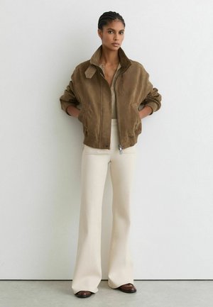 Letitettyä tukkaa oleva nainen pukeutunut ruskeaan mokkatakkiin, beigeen ribbipaitaan, kermaan vivahtaviin leveälahkeisiin housuihin ja ruskeisiin kenkiin, seisoo valkoista seinää vasten.