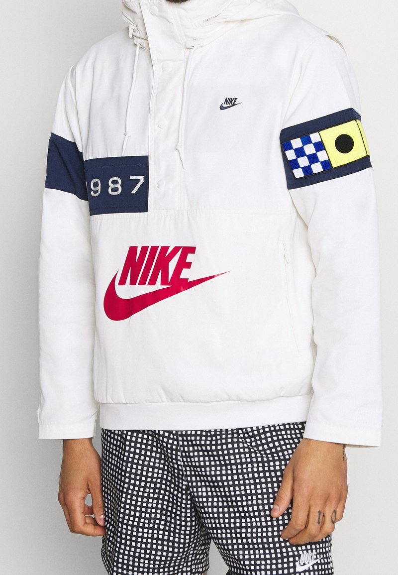 Chubasquero blanco de Nike con un bloque de color azul marino que presenta el año "1987", un gran logo de Nike en rojo y un acento de patrón multicolor en la manga.