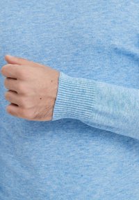 Pull bleu clair avec un tricot texturé, présentant des poignets côtelés. Le tissu semble doux et a un léger motif chiné.