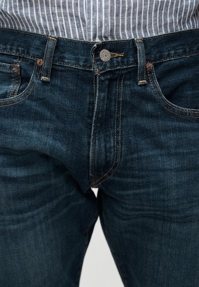 Κοντινό πλάνο ενός ατόμου που φοράει σκούρο μπλε τζιν με πορτοκαλί ραφές και μπλε-λευκό ριγέ πουκάμισο, μαζεμένο μέσα στο παντελόνι.