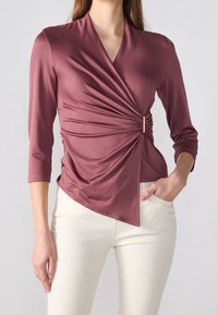 Pitkähihainen, kietaisumallinen paita syvässä mauve-värissä. Ominaisuuksiin kuuluu teksturoitu kangas, sivutaitos yksityiskohtana ja kultainen korostus vyötäröllä.