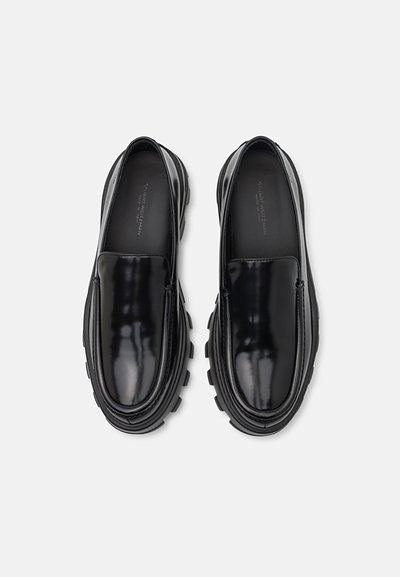 Chaussures noires à enfiler avec une finition brillante, présentant un bout arrondi, une semelle texturée et une semelle extérieure en caoutchouc épaisse et robuste.