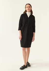 Robe noire à capuche avec des manches trois quarts, dotée d'une poche avant et d'un cordon de serrage élastique. Portée avec des loafers noirs et un col roulé blanc.