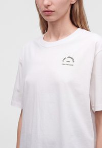 Νεαρή γυναίκα που φοράει ένα λευκό t-shirt με μικρό μαύρο κείμενο "Karl Lagerfeld Paris 21 Rue St-Guillaume" στο αριστερό στήθος.
