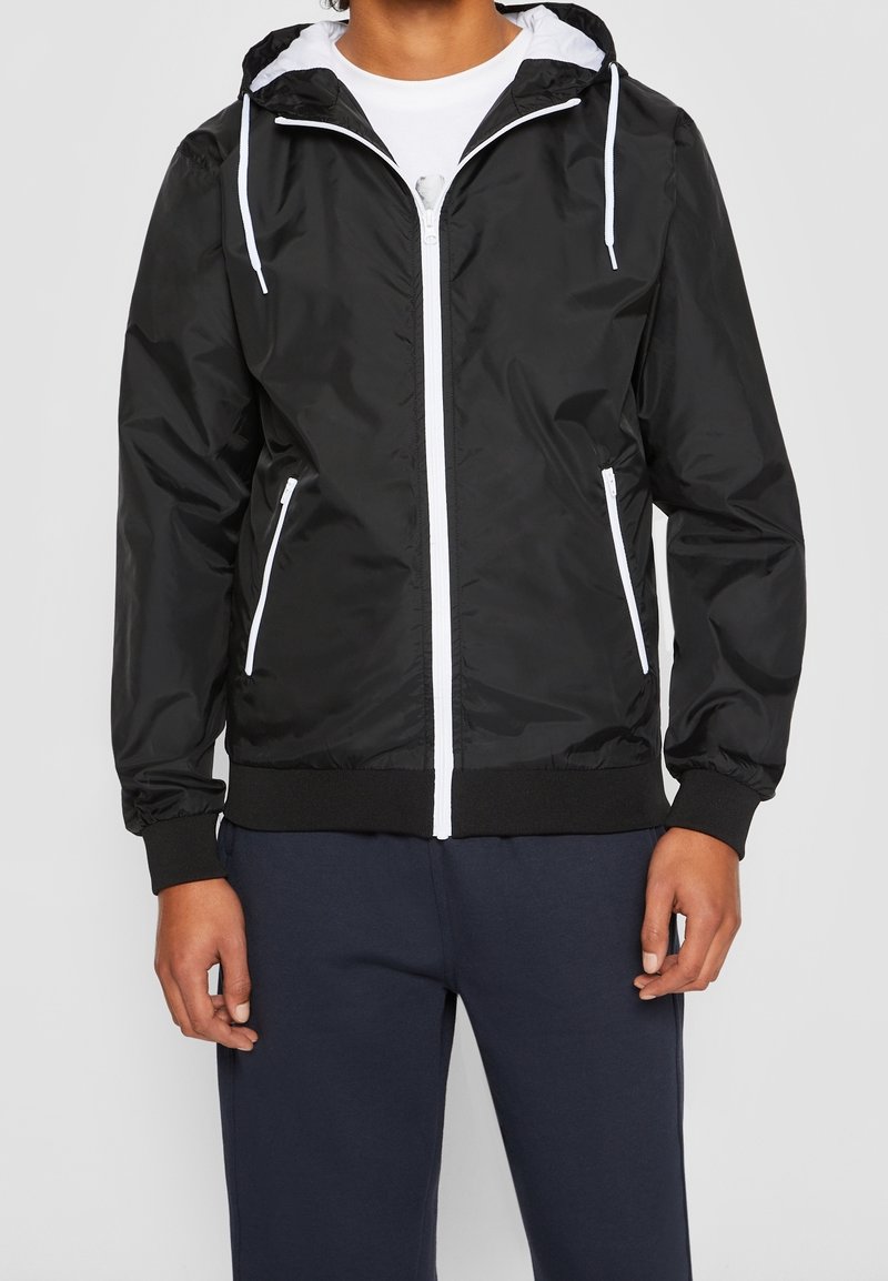Personne portant une veste à capuche noire avec une fermeture éclair blanche et des cordons, associée à un pantalon bleu marine foncé, sur un fond uni.