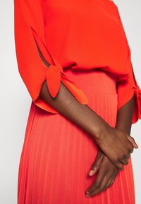 Oranje blouse met strikdetailmouwen, gecombineerd met een plooienrok in dezelfde kleur. Glad textuur, met levendige kleuren en strakke lijnen.
