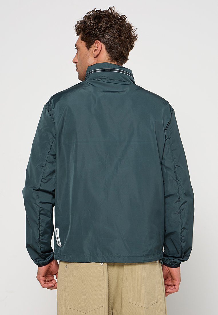 Uomo con capelli ricci che indossa una giacca a vento verde scuro e pantaloni beige, visto di spalle contro uno sfondo neutro.