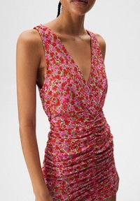 Robe courte fleurie avec un décolleté plongeant en V, mettant en vedette des fleurs roses et rouges sur un tissu texturé. Détails froncés sur les côtés.