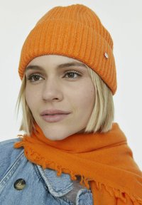 Orange Strickmütze mit vertikaler Rippung und silberner Logoakzent. Kombiniert mit einem orangefarbenen Fransenschal, der über einer Jeansjacke getragen wird.