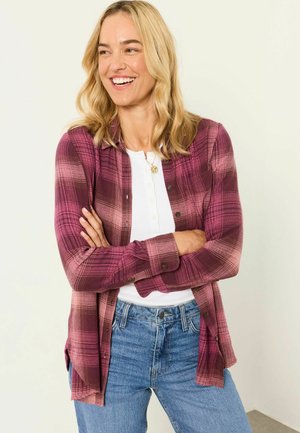 Smiling woman with blonde hair wearing a purple plaid shirt over a white top and blue jeans, arms crossed, standing against a light background.