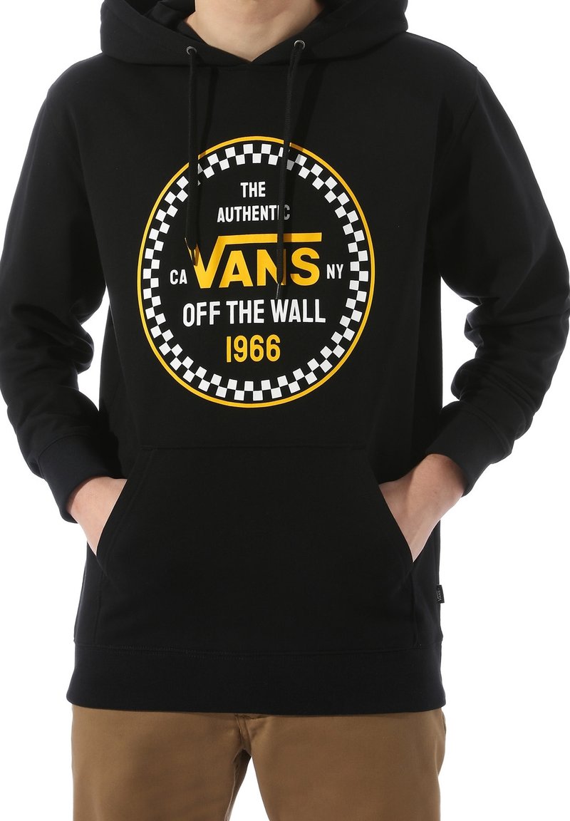 Personne portant un sweat à capuche noir Vans avec un logo circulaire jaune et blanc indiquant « The Authentic Vans Off The Wall 1966. »