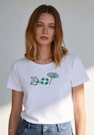 Junge Frau mit schulterlangem hellbraunem Haar, die ein weißes T-Shirt mit grünen Motiven von Strandstuhl, Rettungsring und Sonnenschirm trägt.