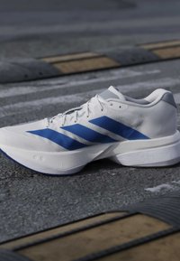 Λευκό αθλητικό παπούτσι με μπλε ρίγες και λεπτομερή σόλα που ξεκουράζεται σε άσφαλτο κοντά σε κίτρινο και μαύρο κορυφαίο εμπόδιο ταχύτητας.