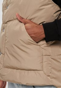Beige polstret vest med en sidelomme og sølvfarvede trykknapper. Har en glat tekstur og en tætsiddende design, vist sammen med en sort ærme.