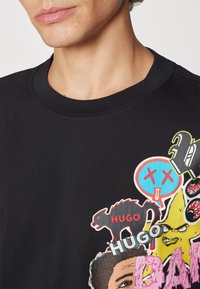 T-shirt in cotone nero con varie stampe grafiche colorate, inclusi volti, testi e simboli, con scollo rotondo e maniche corte.