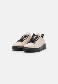 Light beige sneakers with black laces, rounded toe, and textured finish. Features a thick, cushioned black sole and rear pull tab.
