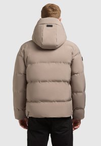 Beige donsjas met een afneembare capuchon, geborduurde textuur en elastische manchetten. Bevat een klein zwart logo bij de hals en een zijrits.