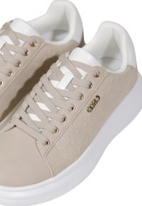 Sneaker beige con una toma superiore testurizzata, accenti bianchi, lacci piatti e occhielli in tonalità oro. Presenta una placca con logo sul lato e una suola spessa di colore bianco.