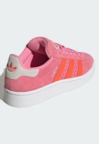 Sapatilha de camurça rosa com riscas laranja, sola branca e detalhe cinza no calcanhar. Apresenta logótipo e atacadores texturizados. Design casual para uso diário.