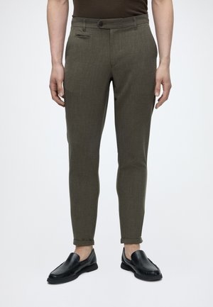 Homme portant un pantalon slim vert olive avec des ourlets retroussés et des mocassins en cuir noirs, debout sur un fond blanc.