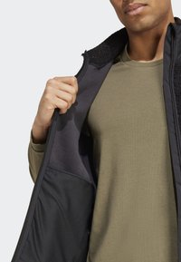 Schwarze, fleecegefütterte Jacke mit Reißverschluss vorne, die ein grünes Langarmshirt darunter zeigt. Sie hat strukturierte Akzente und eine innere Netz-Innentasche.