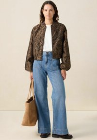 Veste bomber imprimée léopard marron avec fermeture éclair à l'avant, associée à un jean à jambes larges bleu clair. Les accessoires comprennent un sac fourre-tout en suède taupe.