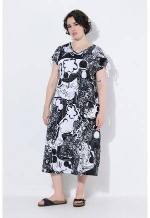 Frau mit kurzen lockigen Haaren, die ein schwarz-weiß geblümtes Midikleid und schwarze Slip-On-Sandalen trägt, steht vor einem weißen Hintergrund.