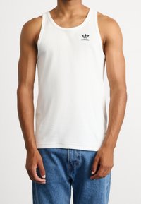 Camiseta de tirantes blanca con textura de canales, con un escote de cuello redondo y un pequeño logo negro de Adidas en el lado izquierdo del pecho, combinada con unos jeans de denim azules.