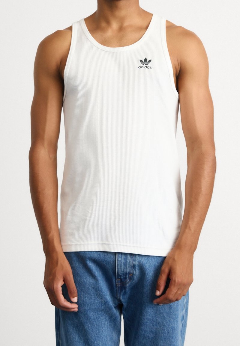 Camiseta de tirantes blanca con textura de canales, con un escote de cuello redondo y un pequeño logo negro de Adidas en el lado izquierdo del pecho, combinada con unos jeans de denim azules.