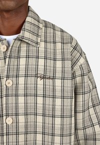 Kockás, bézs és fekete gombos kabát. Hajtóka, bézs gombok és egy diszkrét hímzett logó található a mellkason. Texturált anyag.