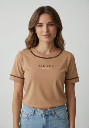 Giovane donna con capelli castano ondulati indossa una t-shirt beige "YES ZEE Natural Feeling" con rifiniture scure e jeans azzurri, in piedi davanti a uno sfondo semplice.