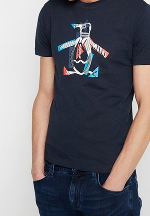Homme portant un t-shirt bleu marine foncé avec un motif abstrait de pingouin multicolore, mains décontractées dans les poches, associé à un jean bleu foncé.