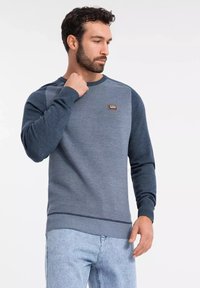 Suéter azul marino con puños y dobladillo acanalados, que presenta un patrón texturizado y una pequeña placa de logotipo rectangular en el pecho.