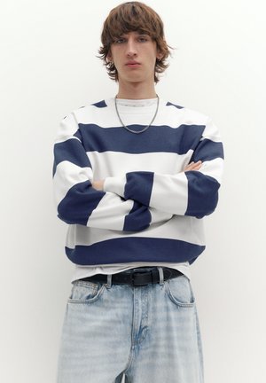 Mlada oseba s srednje dolgimi rjavimi lasmi, oblečena v belo in mornarsko modro črtasto majico, svetlo modre kavbojke in srebrno verižico okoli vratu, stoji z zamašenimi rokami.