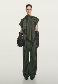 Kobieta w ciemnozielonej asymetrycznej bluzce i szerokich spodniach, nosząca długie czarne skórzane rękawiczki i trzymająca czarną skórzaną torebkę kopertową.