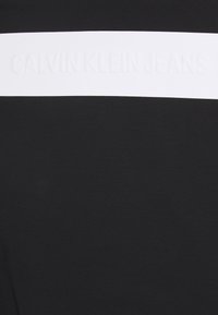Tissu en coton noir présentant une large bande horizontale blanche avec le texte embossé "CALVIN KLEIN JEANS." Le design est minimaliste et contemporain.