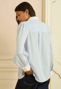 Vaaleansininen ja valkoraidallinen paita, jossa kauluksellinen pääntie, liehukkeet hihansuissa ja pitsireunaiset kalvosimet. Rentoa istuvuutta ja pehmeä kangaspinta.