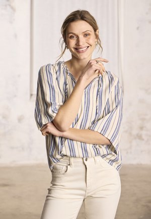 Mladá žena sa usmieva, nosí krémové nohavice a voľnú bielu blúzku s vertikálnymi modrými pruhmi, stojí so založenými rukami v svetlej miestnosti.