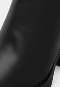 Crna kožna gležnjača s sjajnim završetkom, okruglim vrhom, glatkom teksturom i niskom slojevitom potpeticom.