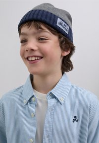 Harmaa ja laivastonsininen raidallinen neulottu pipo, jossa on lappu, yhdistetty vaaleansiniseen ja valkoiseen raidalliseen nappilistalliseen paitaan, jossa on pieni pääkallokuvio.