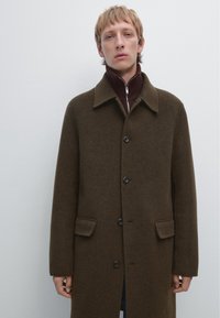 Manteau en laine marron avec un col à revers, fermeture à boutons et deux poches avant. Présente un pull bordeaux superposé en dessous.